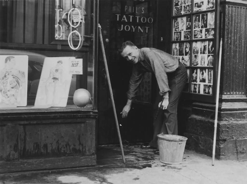 A man crouches and washes the sidewalk outside a tattoo shop called “Phil’s Tattoo Joynt.” There’s a bucket, mop, and tattoo art displayed in the shop window and on the wall.