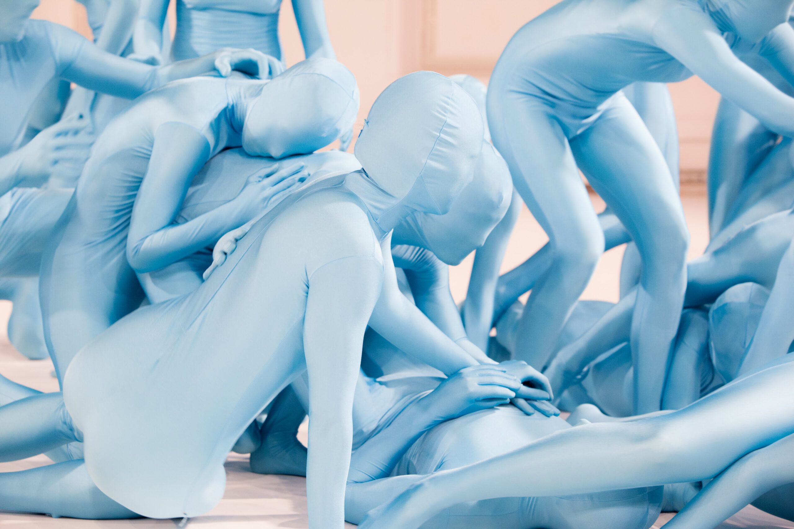 A group of people wearing full-body blue suits interact closely, overlapping and supporting each other in dynamic poses on a light background.