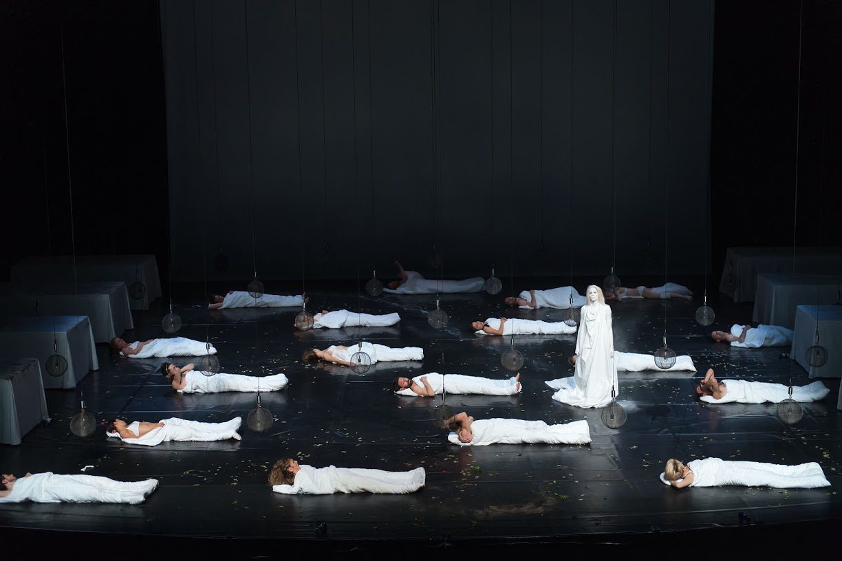 A stage scene with one person standing in white, surrounded by many others lying on the floor in white costumes, under dim lighting, portraying a dramatic or theatrical performance.