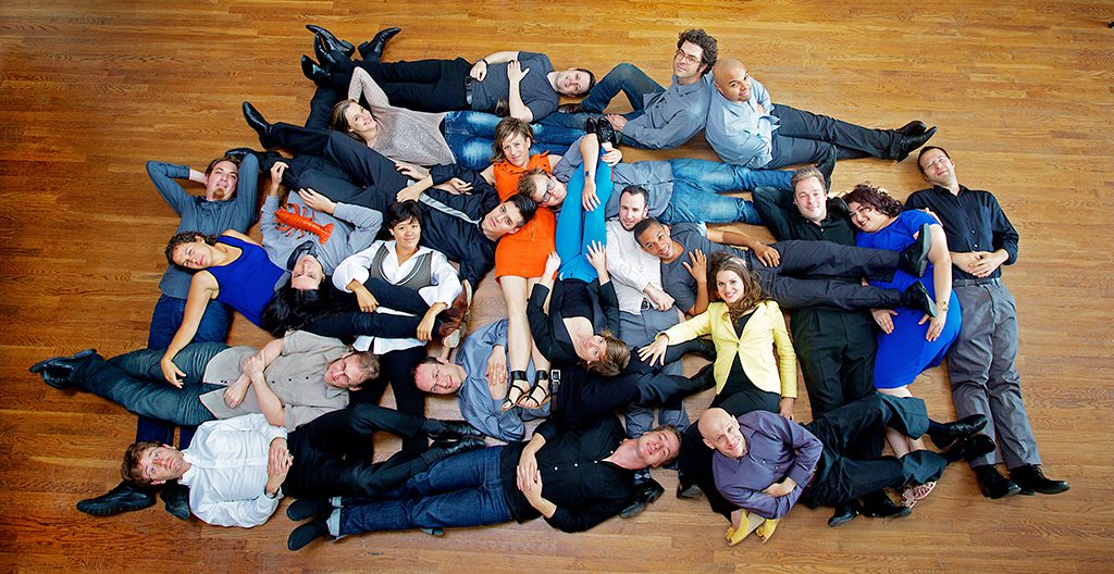 Thirty people lie on a wooden floor, overlapping and arranged in a loose circle, some in bright clothing and others in darker tones, creating a visually striking, casual pattern with playful, relaxed expressions and poses.