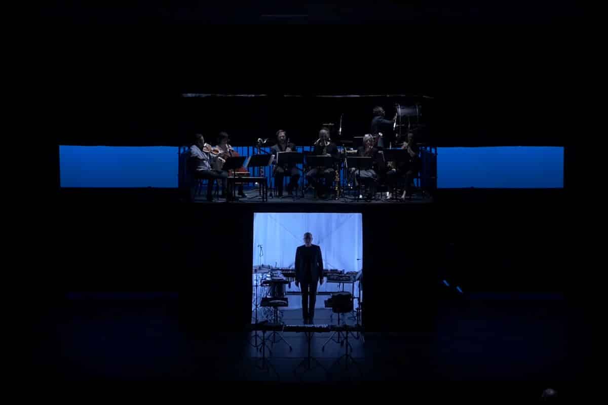 A dimly lit stage shows musicians seated above, playing instruments, while a person stands alone in a lit area below, surrounded by percussion instruments.