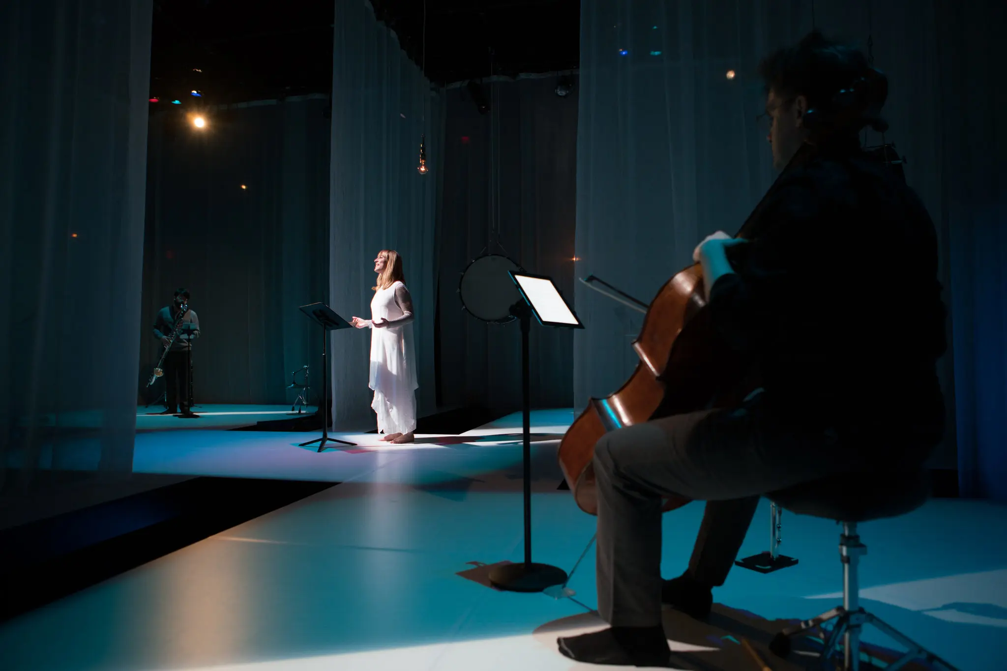 A cellist plays in the foreground while a woman stands singing at a music stand under stage lights, with a saxophonist visible in the background; sheer curtains hang around the stage.