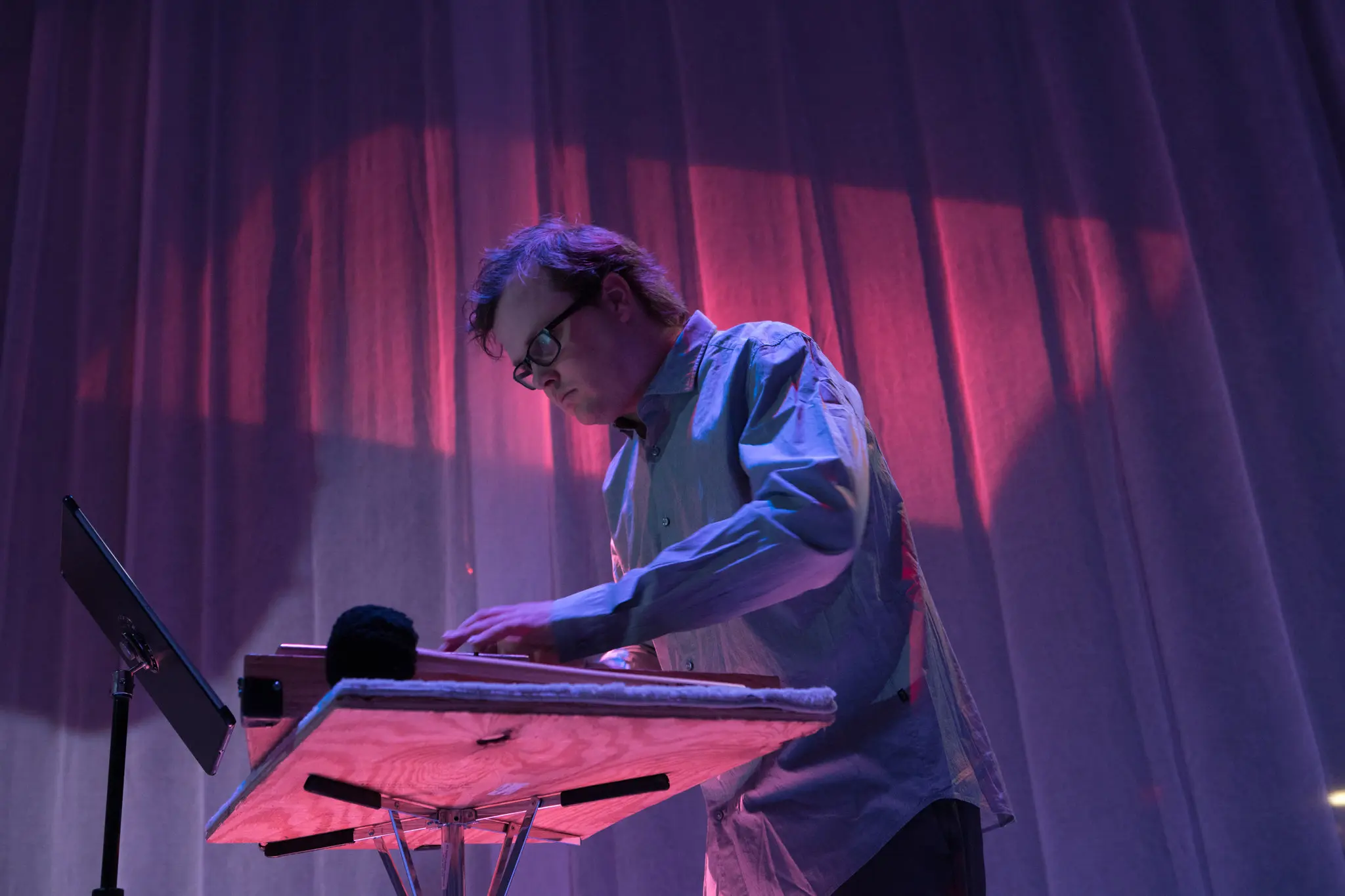 A person in glasses plays an electronic instrument on stage under purple and pink lighting, with a music stand and curtains in the background.