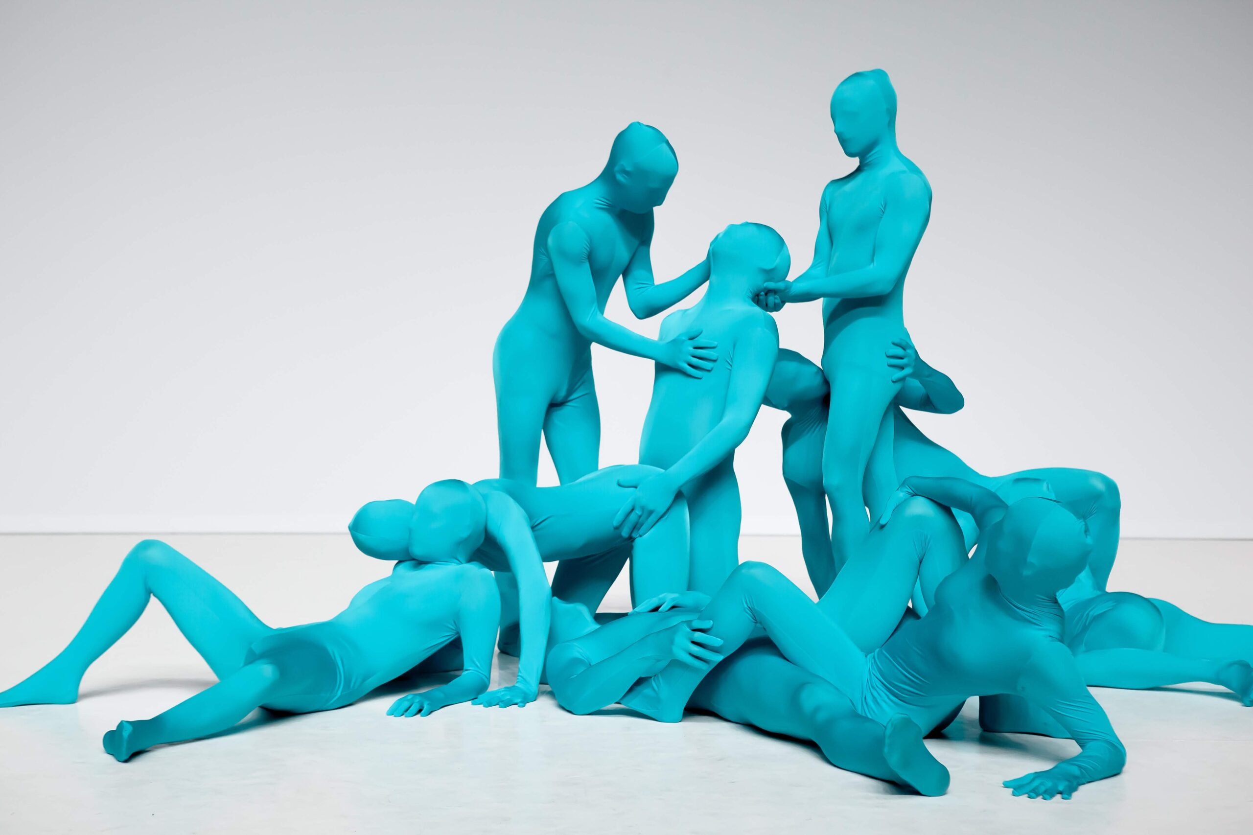 Nine people in tight, full-body turquoise suits pose in an abstract, interconnected arrangement against a white background. Their faces and features are completely obscured by the suits.