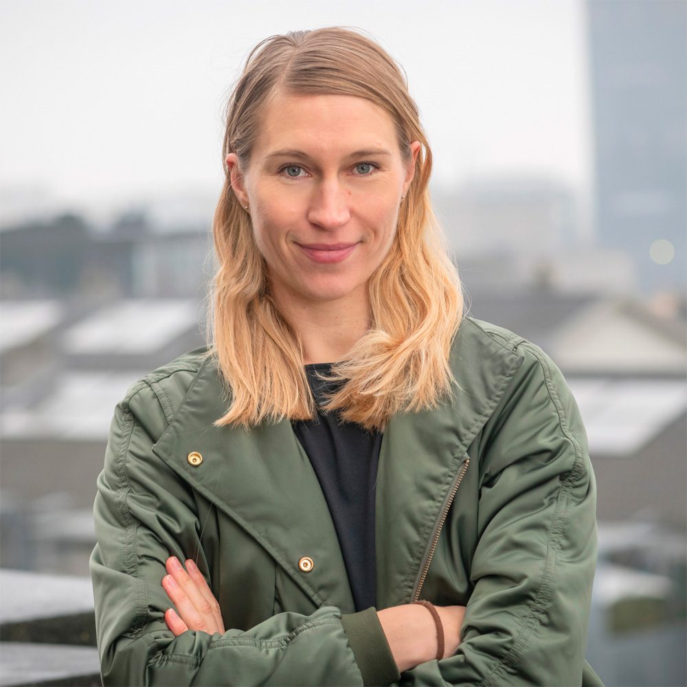 A woman with blonde hair in a green jacket stands outdoors with her arms crossed, smiling slightly. The background is blurred buildings and a cloudy sky.