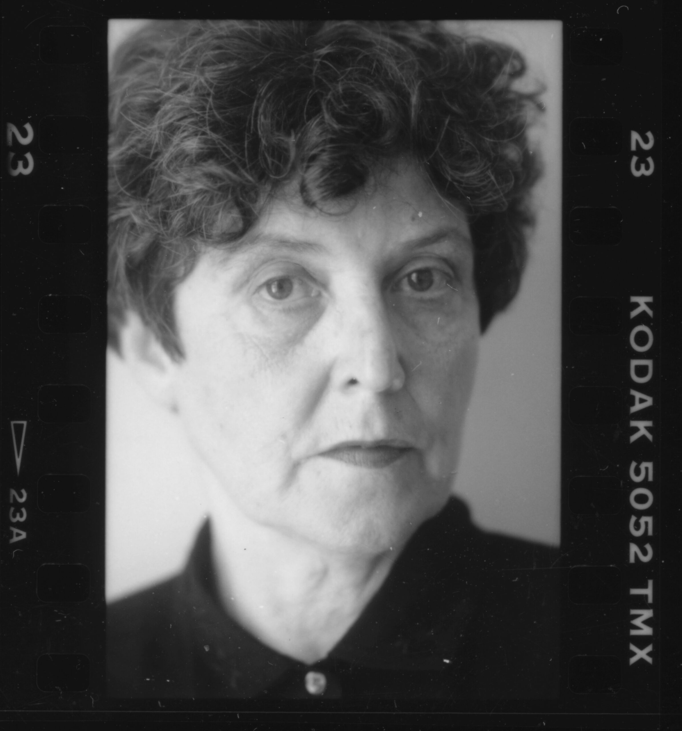 Black and white portrait of a person with short, curly hair, wearing a dark shirt, looking directly at the camera with a neutral expression. The image has a film border labeled “KODAK 5052 TMX.”.