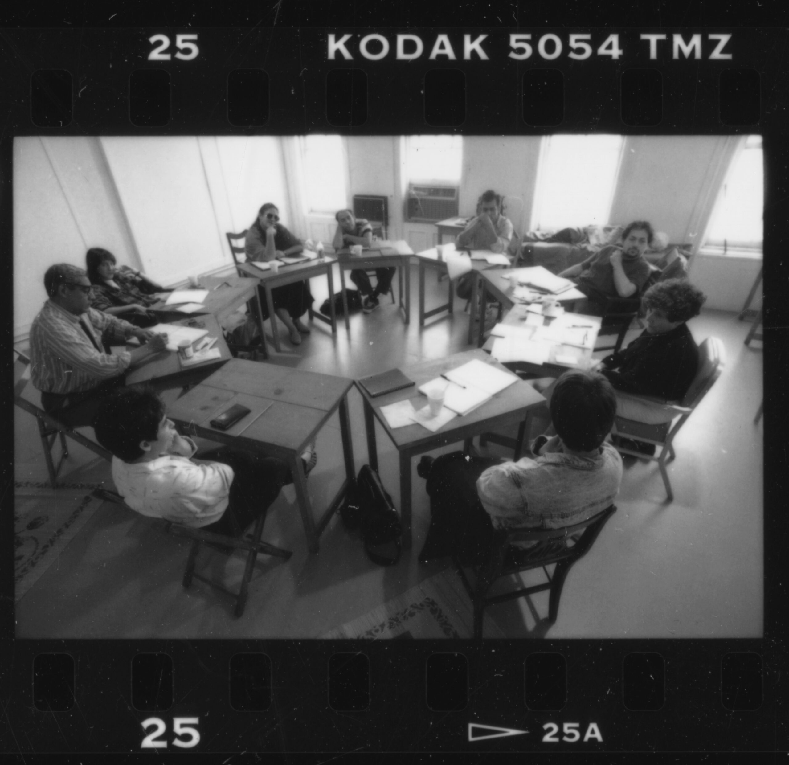 Black and white photo of nine people sitting around a cluster of tables in a meeting room, engaged in discussion with papers and notebooks on the desks. Image has a film negative border marked “KODAK 5054 TMZ.”.