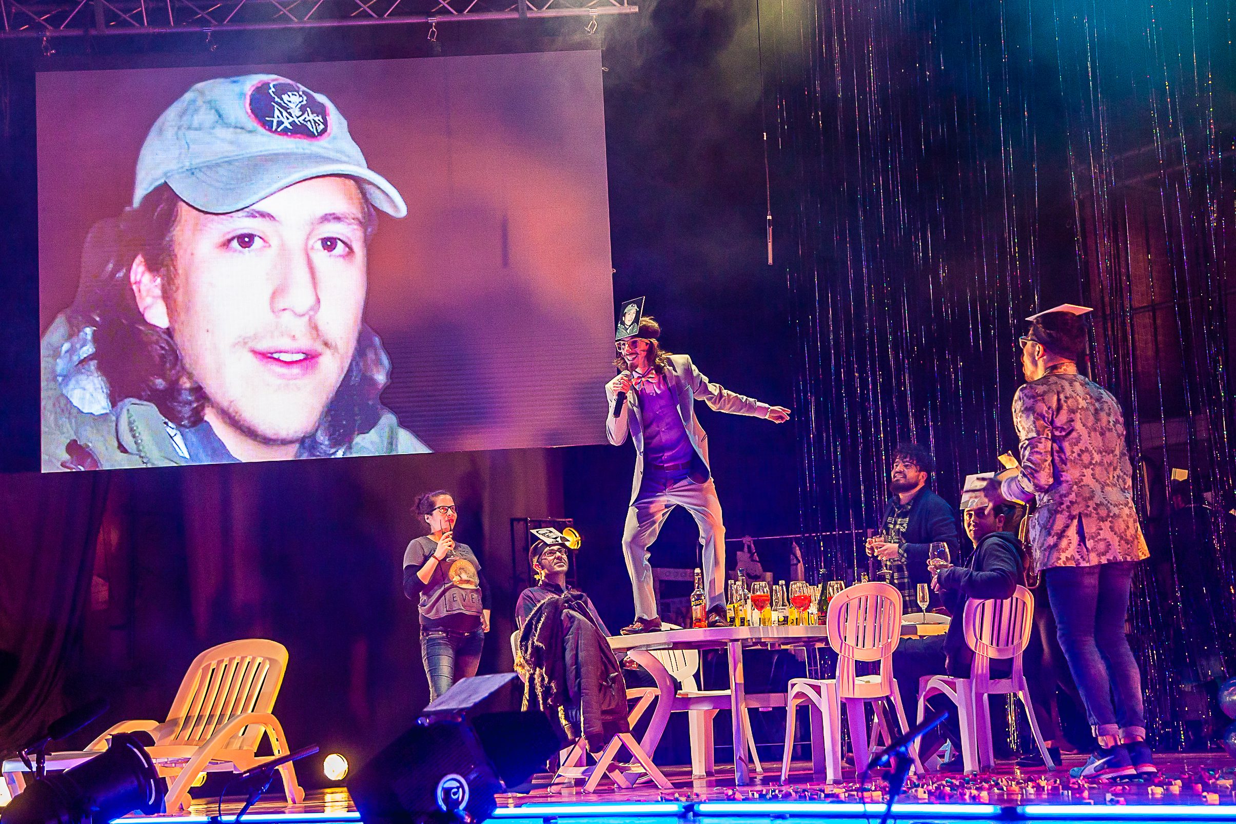 A performer stands on a table during a vibrant stage show, while others sit or stand nearby. A large projected image of a man’s face with a cap is displayed in the background. Bright, colorful lighting fills the scene.