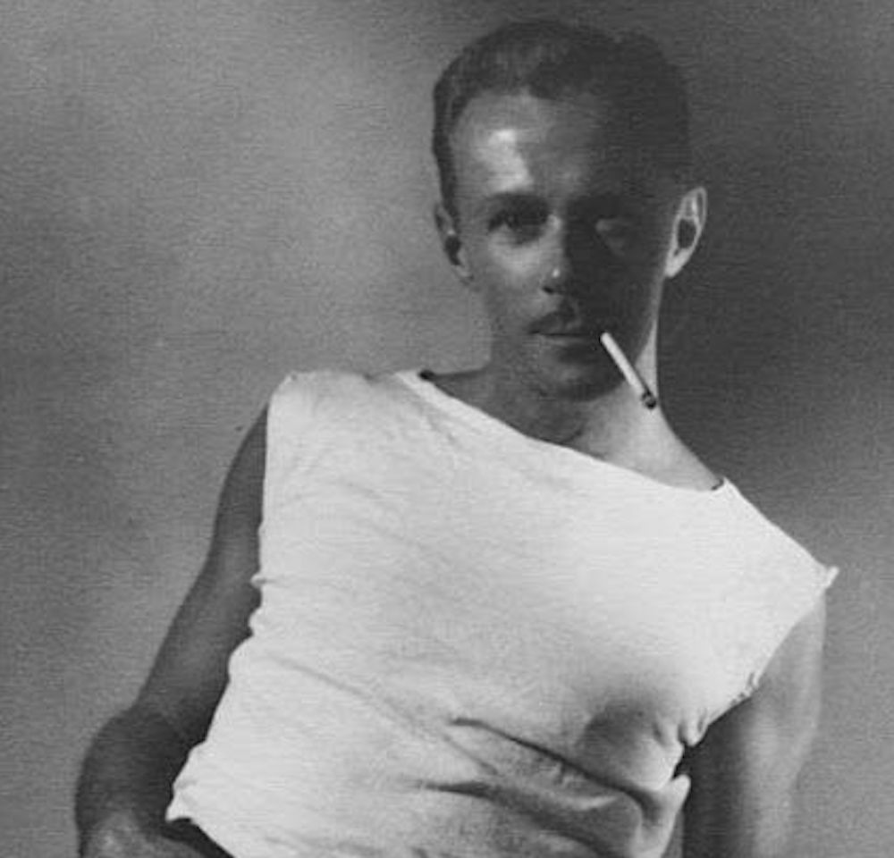 Black and white photo of a man in a white sleeveless shirt, leaning slightly with a cigarette in his mouth, looking toward the camera with a shadowy background.