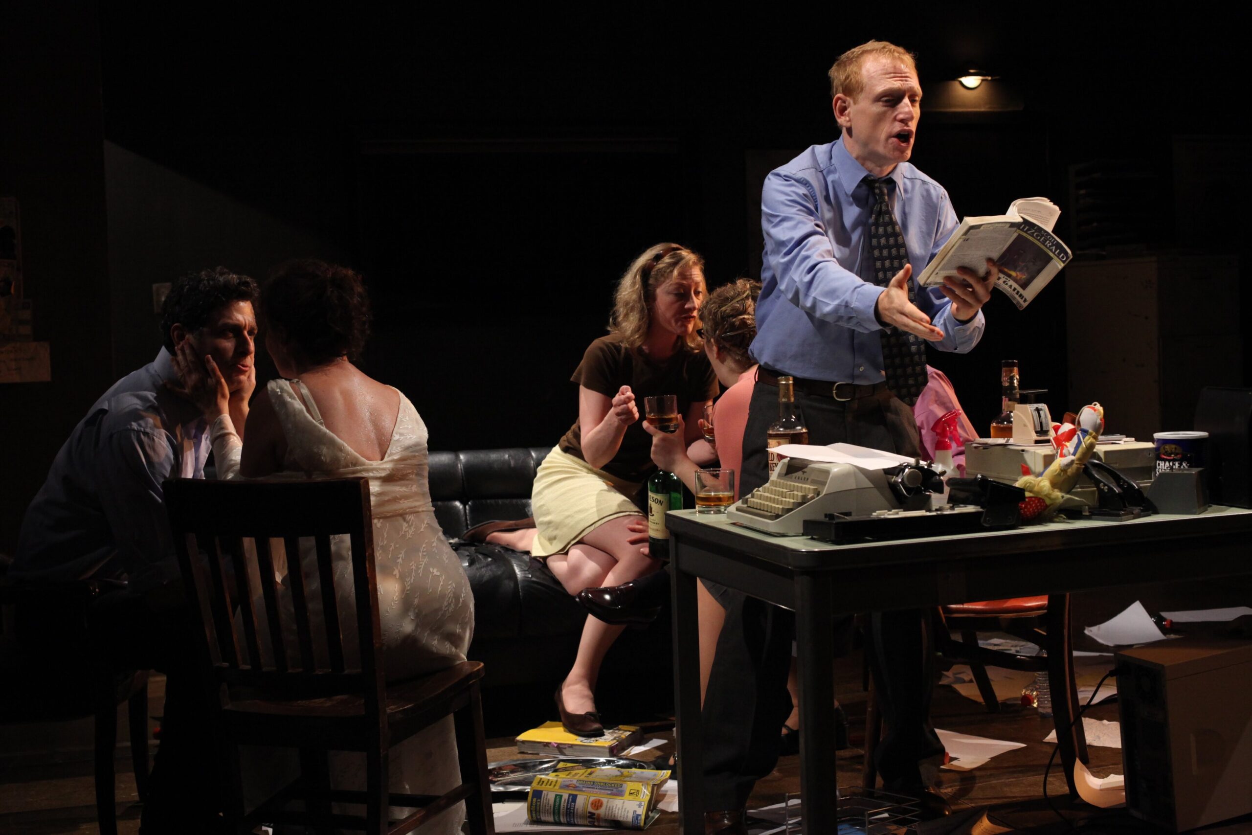 Six adults talk and drink in a dimly lit room; one man stands at a cluttered desk, reading from a book, while others sit on a couch and chairs, engaged in conversation.