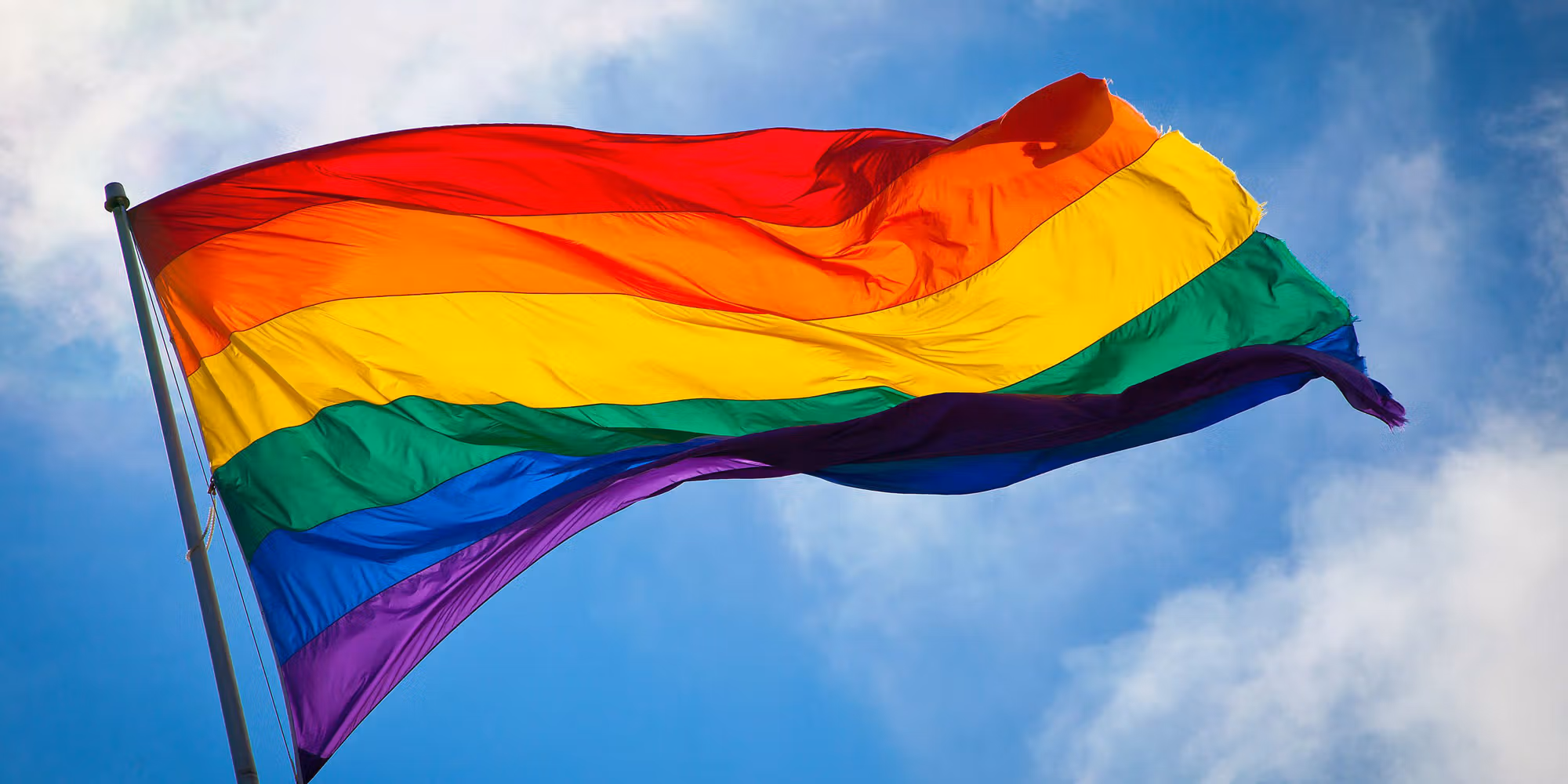 A rainbow-colored pride flag waves against a blue sky with some clouds.