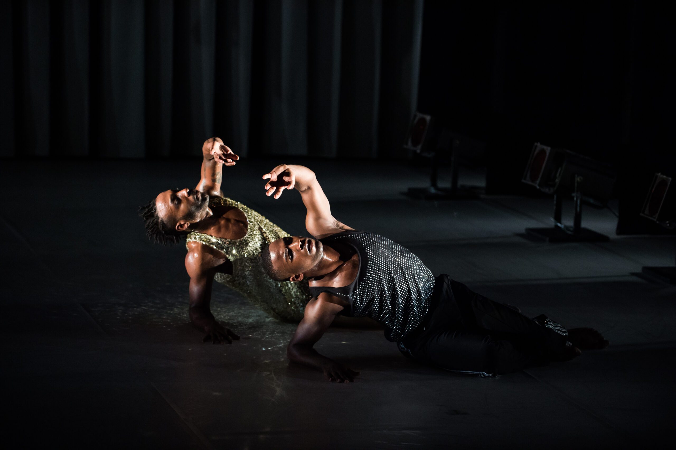 Two dancers in glittery costumes lie side by side on stage, raising one arm each in expressive poses under dramatic lighting, with a dark background and focused spotlights highlighting their movements.