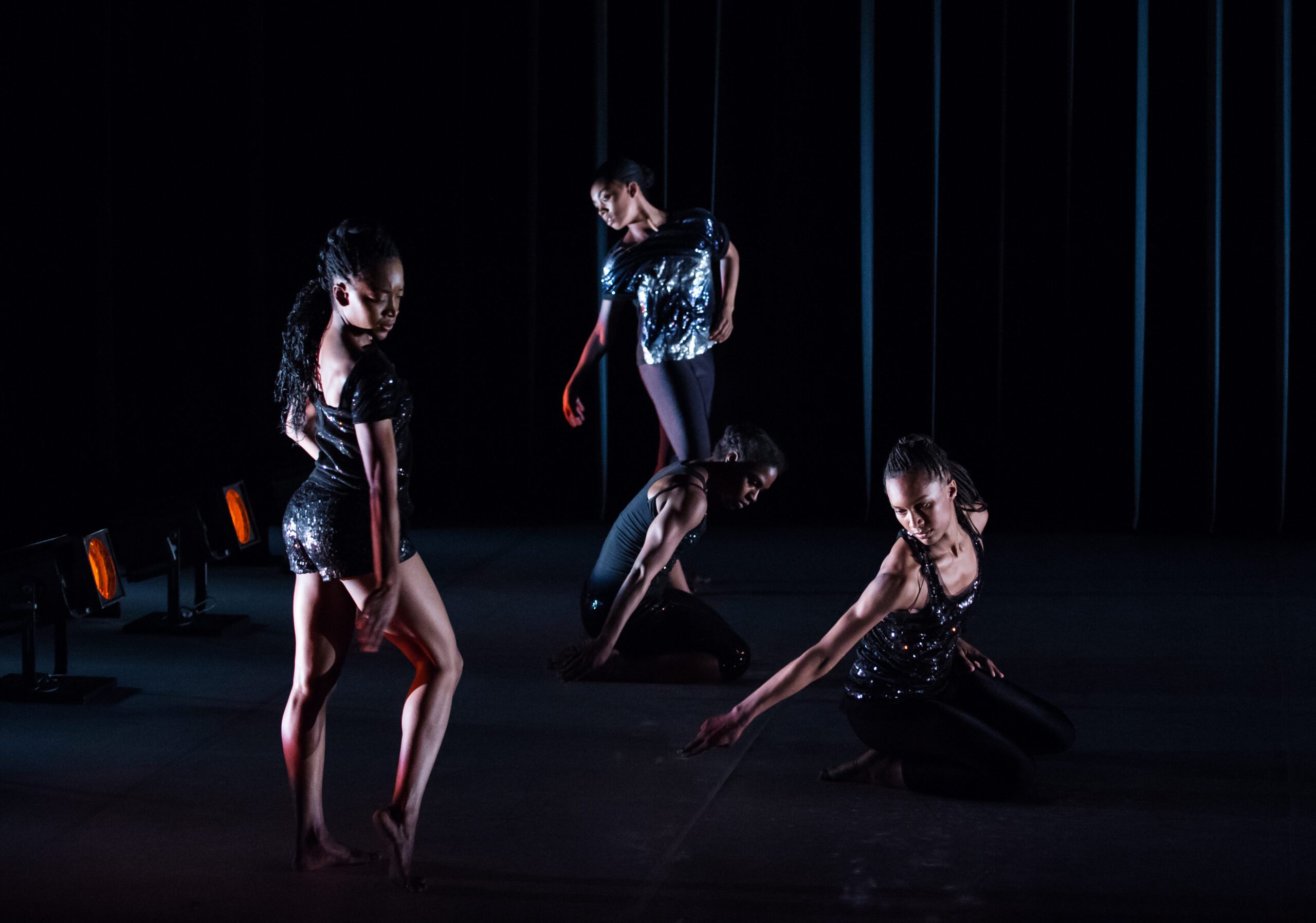 Four dancers in dark, sparkly outfits perform on a dimly lit stage, striking expressive poses with dramatic lighting and shadows enhancing the scene.