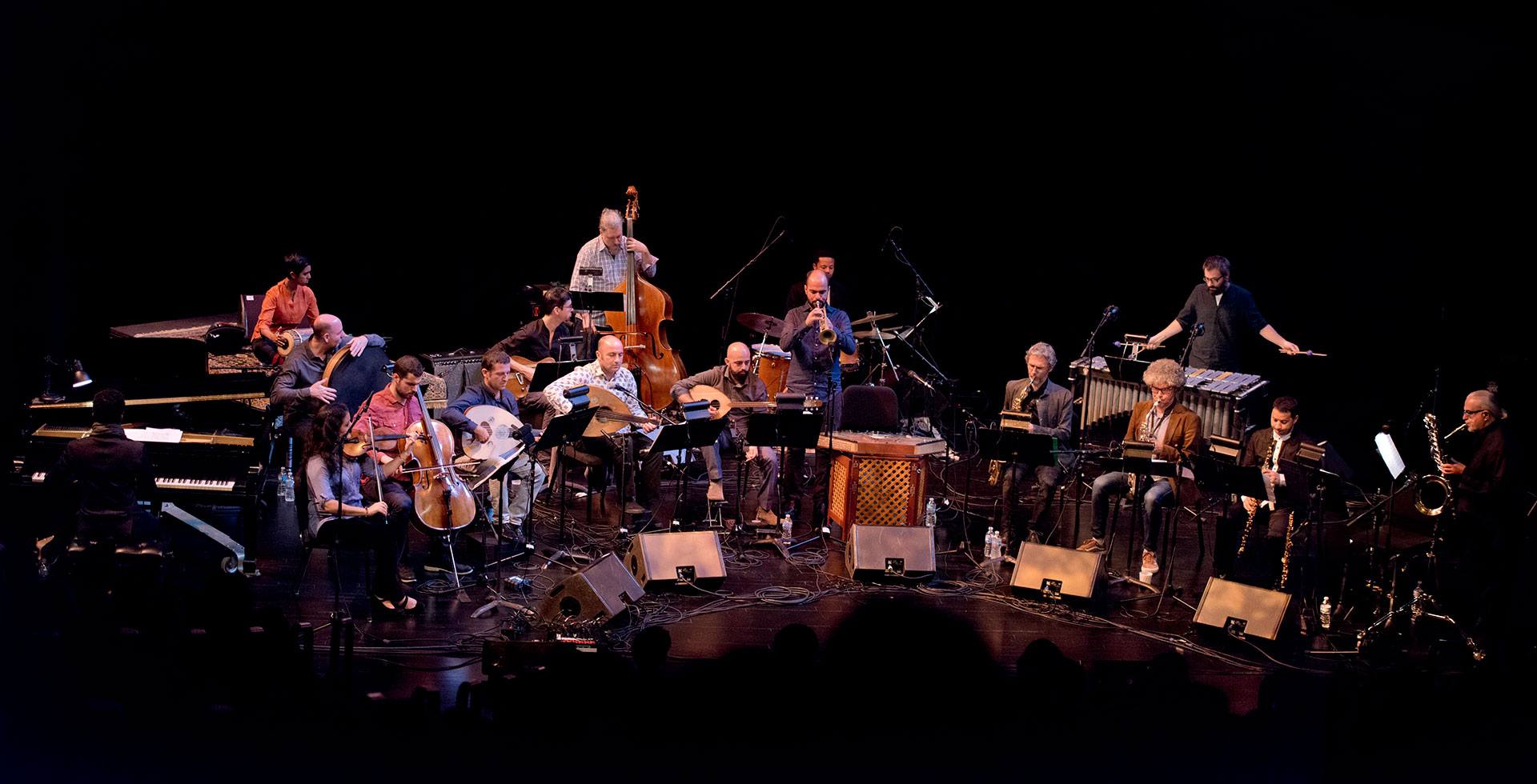 A large ensemble of musicians plays various instruments, including strings, brass, woodwinds, percussion, and piano, on a dimly lit stage with sheet music stands and microphones.