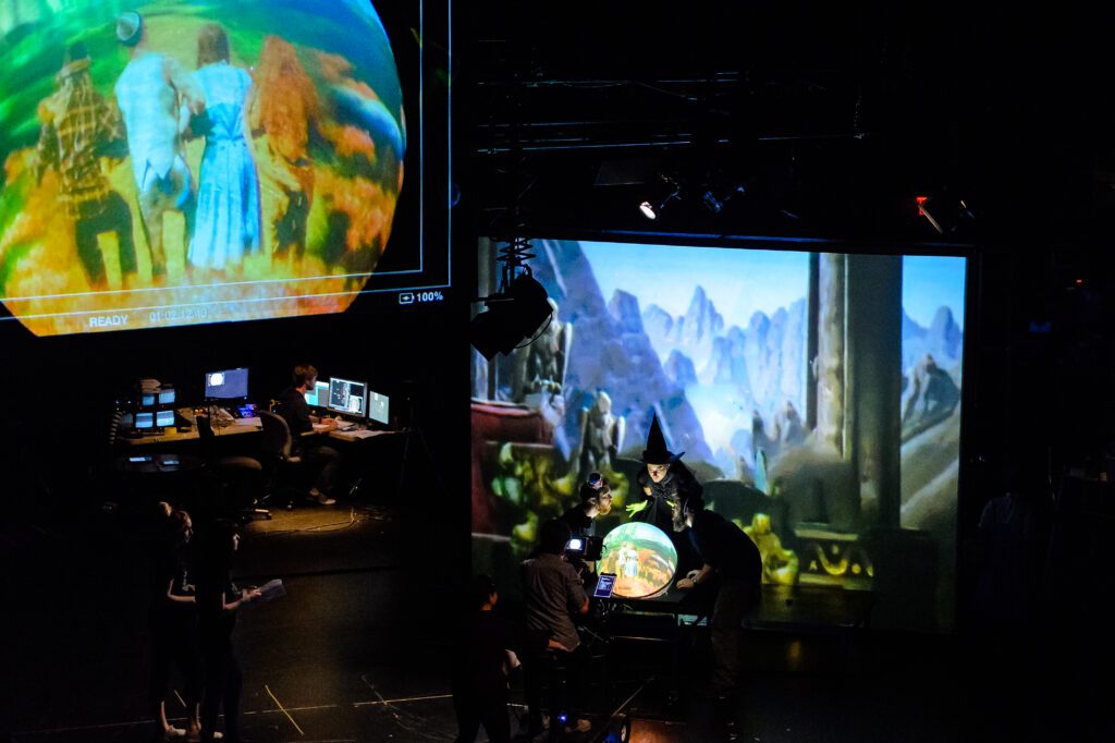 A dark theater shows people operating computers, while a large screen displays a scene from “The Wizard of Oz” with characters and a witch looking into a crystal ball.