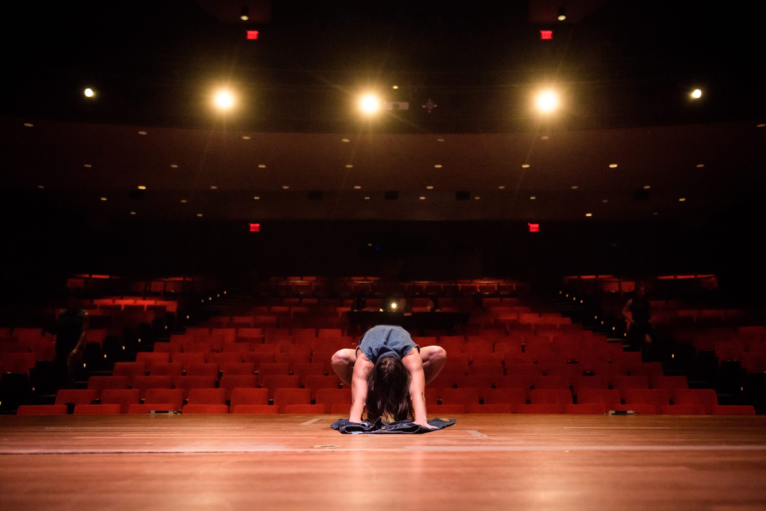A person on stage bends forward, touching the floor with their head, facing an empty, dimly lit theater with rows of red seats and bright lights overhead.