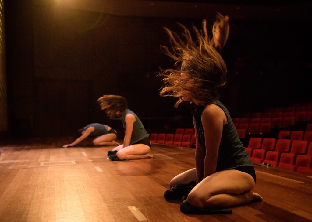 Three dancers perform on a dimly lit stage, kneeling and swinging their hair dramatically. The theater seats are mostly empty in the background.
