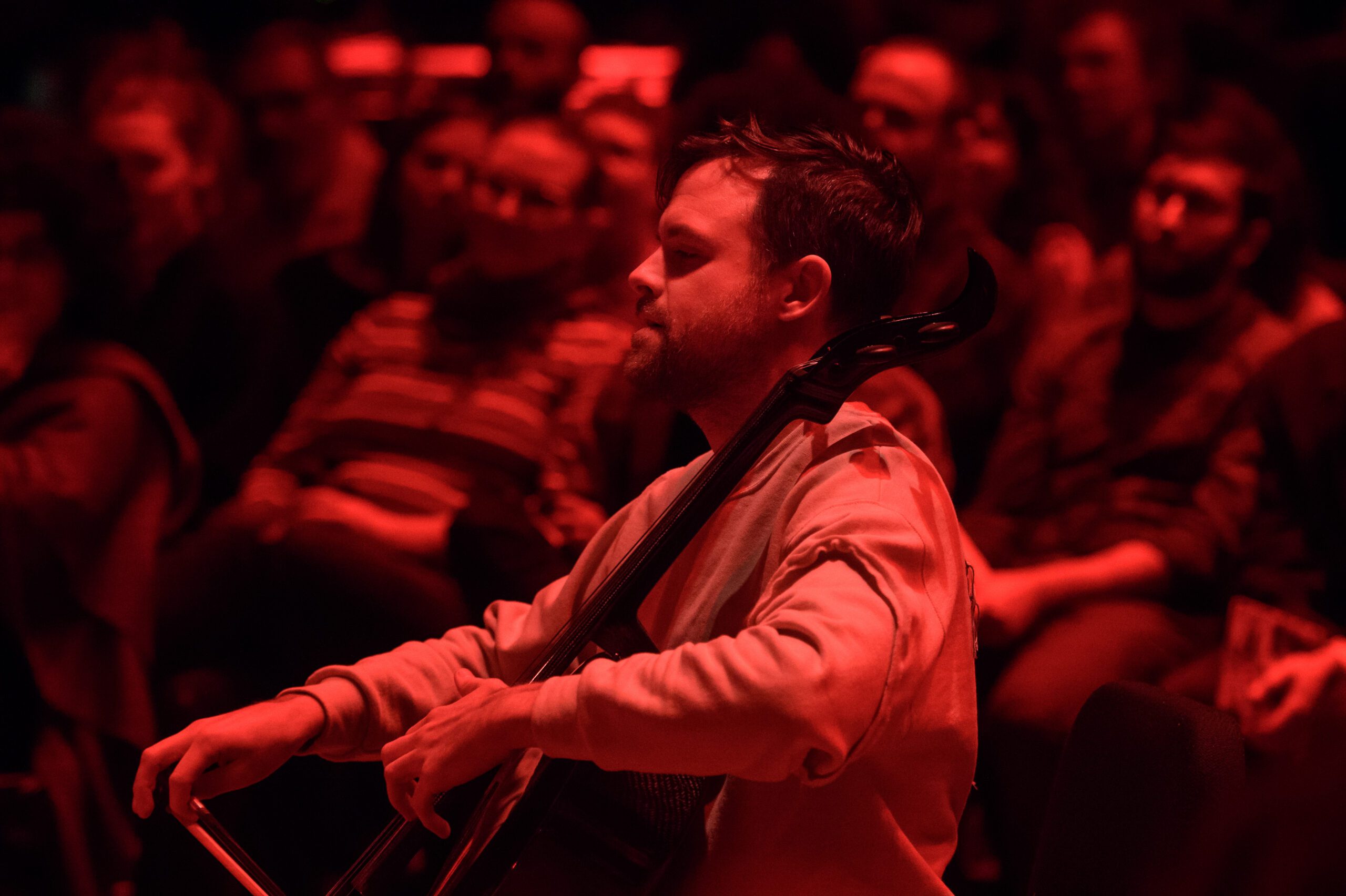 A cellist, illuminated by red stage lighting, plays in front of an audience seated in the background. The atmosphere is focused and intimate.