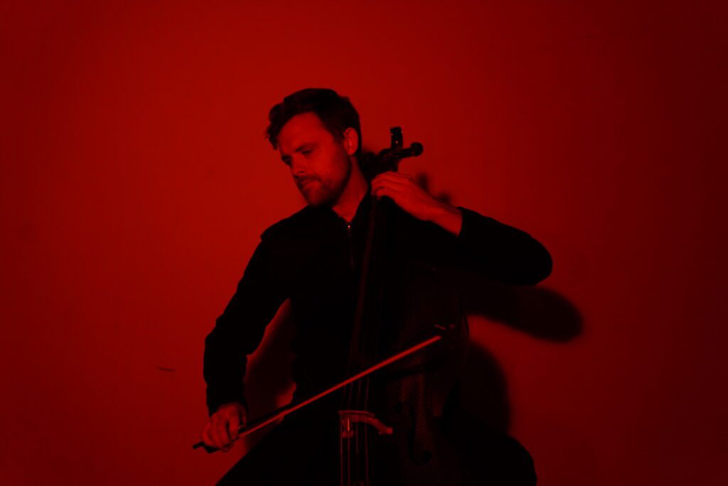 A person plays a cello against a plain red background, lit by dramatic red lighting. They wear dark clothing and appear focused on the music.