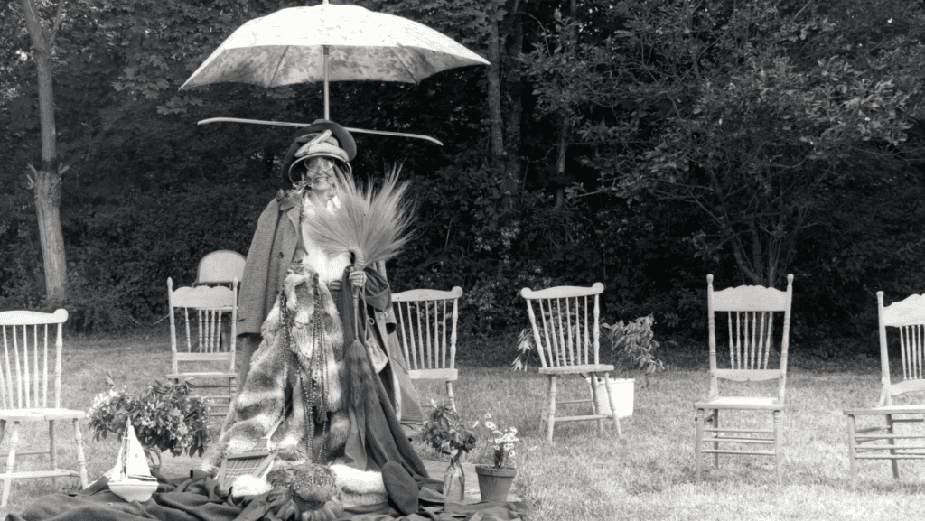 A person stands outdoors in vintage clothing and hats, holding large grass plumes, with an open umbrella overhead. They are surrounded by empty wooden chairs, potted plants, and trees in the background. The image is black and white.