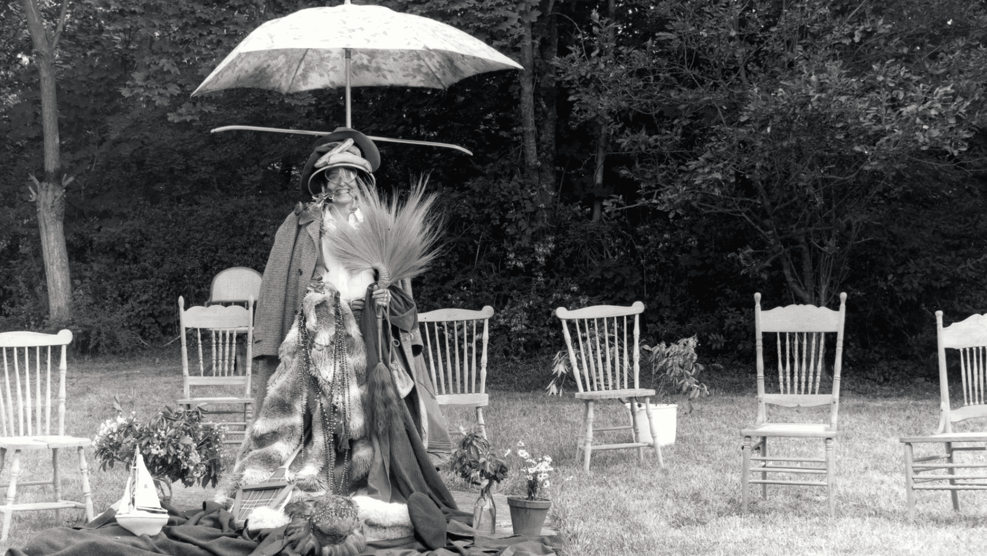 A person stands outdoors in vintage clothing and hats, holding large grass plumes, with an open umbrella overhead. They are surrounded by empty wooden chairs, potted plants, and trees in the background. The image is black and white.