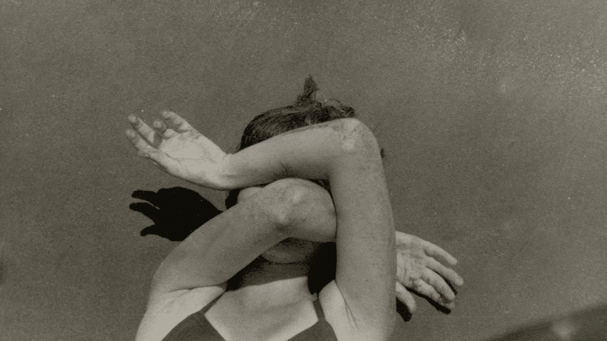 A person in a swimsuit sits with both arms crossed over their face, partially blocking their head and casting dramatic shadows on a textured wall behind them. The photo is in black and white.