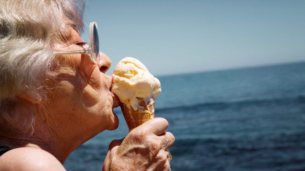 An elderly woman with gray hair and glasses enjoys eating an ice cream cone by the ocean on a sunny day.