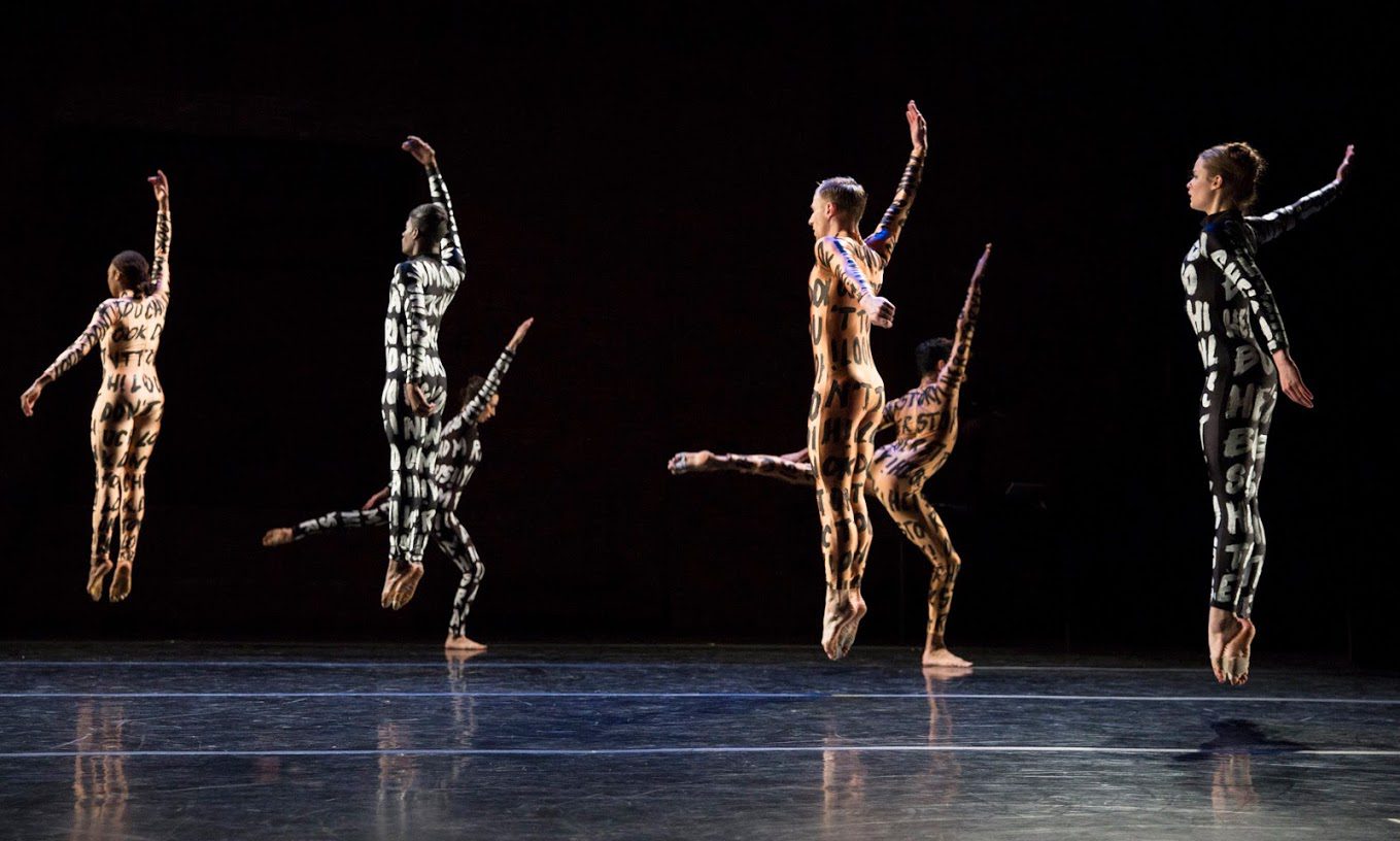 Six dancers in patterned bodysuits leap mid-air on a dimly lit stage, their arms and legs extended in unison, creating a dynamic and synchronized performance.