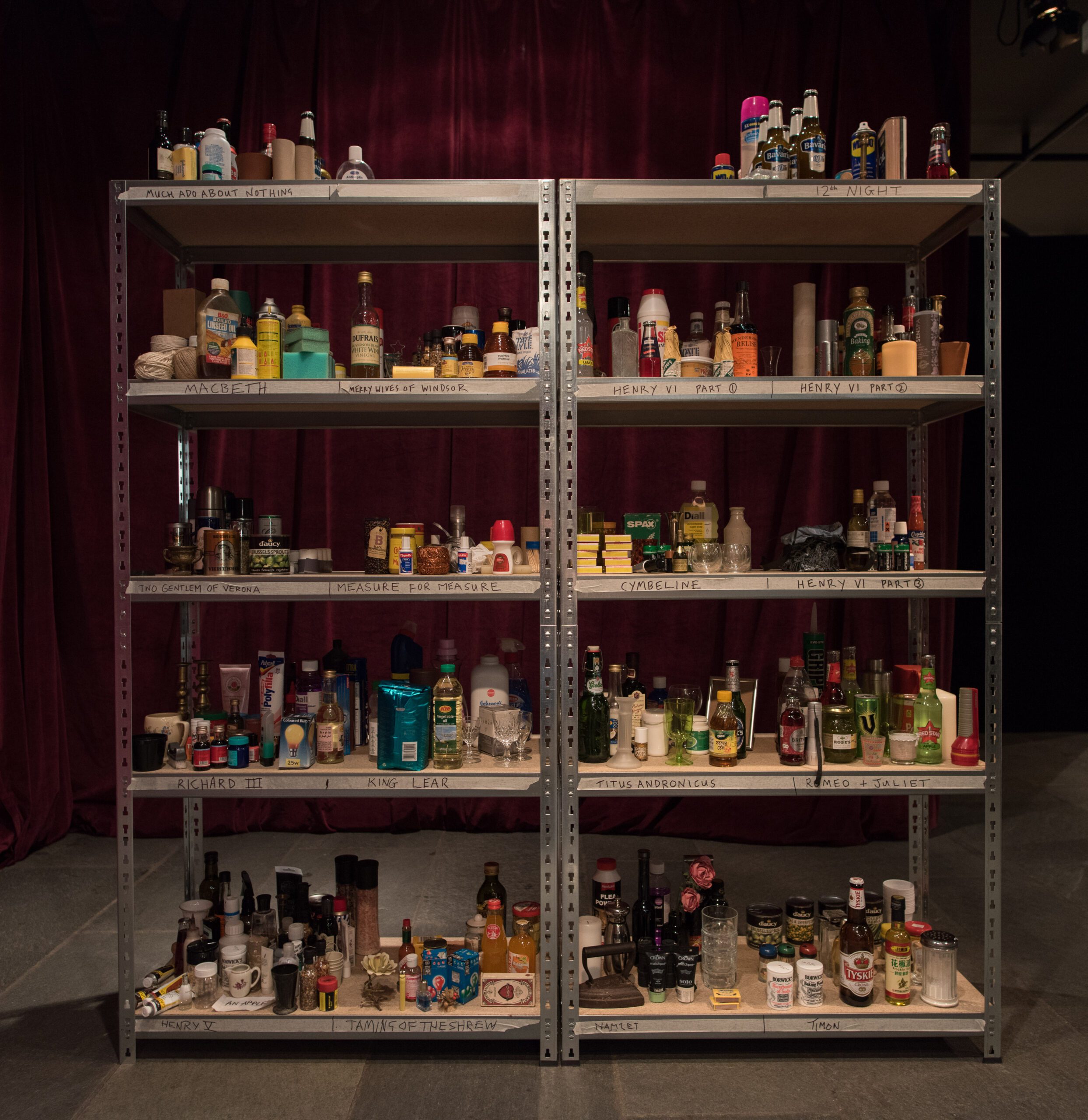 A metal shelving unit holds assorted bottles, cans, household products, and personal items, organized on five shelves, against a backdrop of maroon curtains.