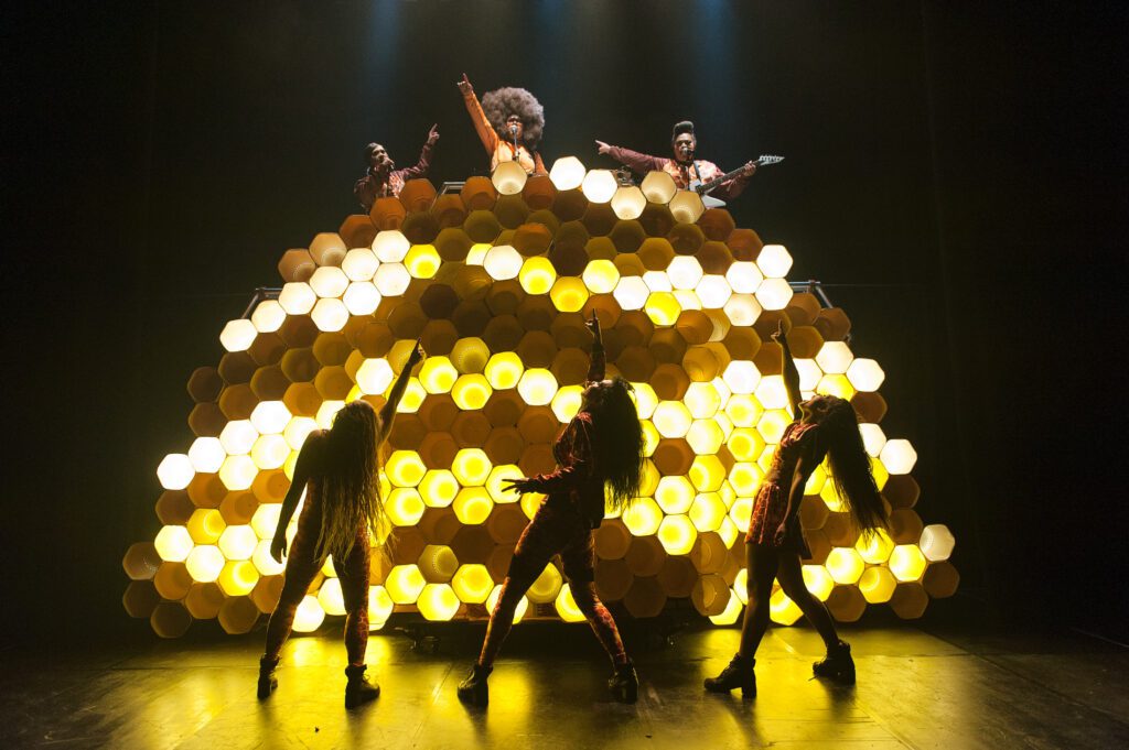 Four performers pose in front of a glowing, honeycomb-shaped set, while three others stand on top, dramatically lit in colorful costumes. The stage is dark except for the illuminated set.