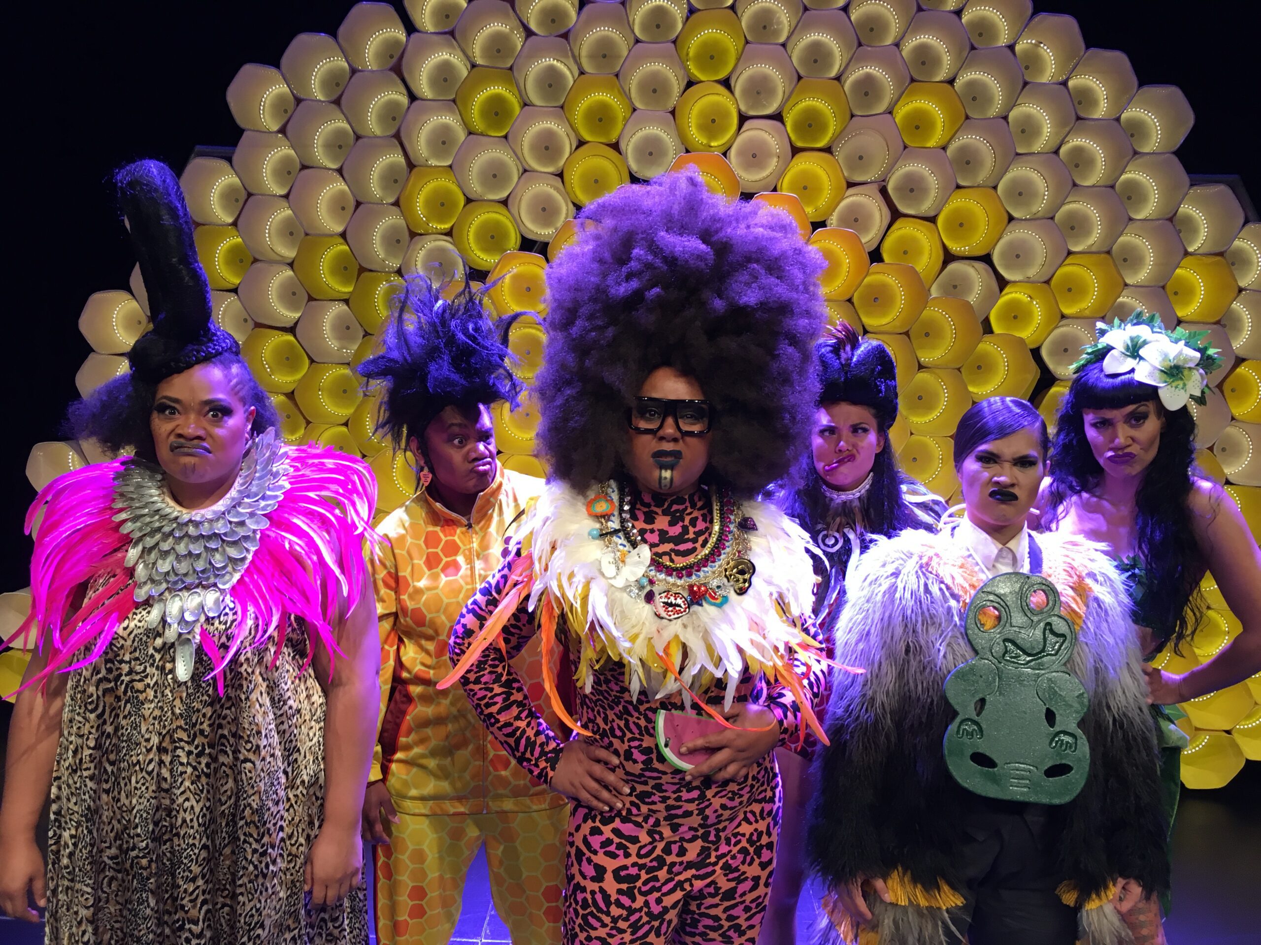 Six people in colorful, extravagant costumes stand in front of a yellow, flower-like backdrop, each with dramatic hair, makeup, and accessories, appearing ready for a performance.