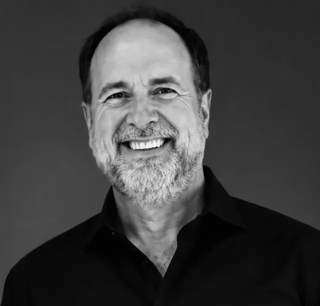 A middle-aged man with a beard and short hair smiles warmly at the camera. He is wearing a dark shirt and the background is plain, creating a professional and friendly portrait in black and white.