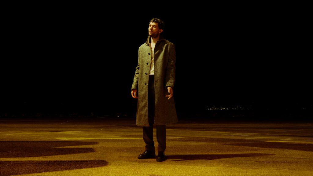 A man in a long gray coat stands alone on an empty, dimly lit pavement at night, surrounded by darkness with faint city lights in the background.