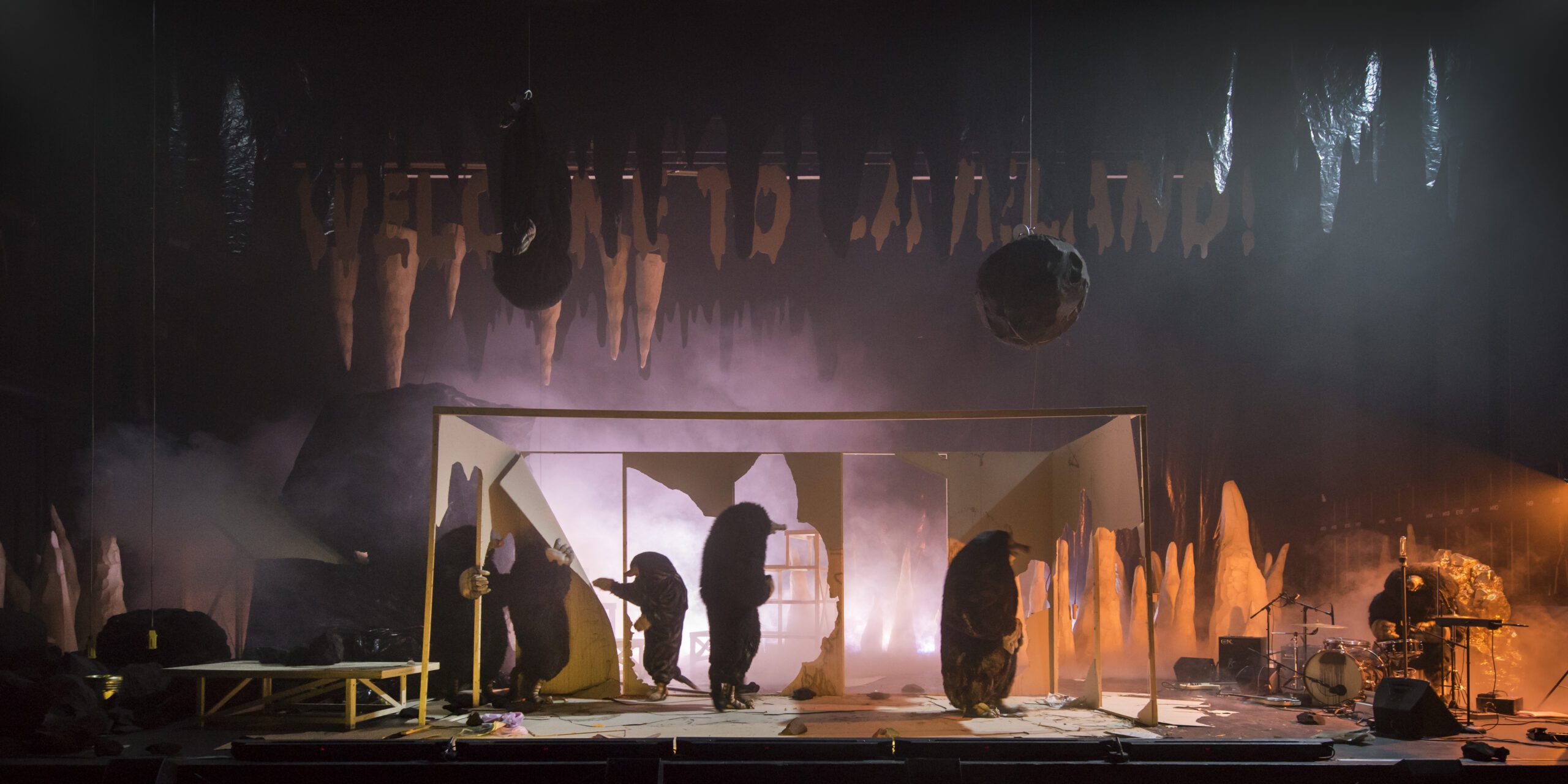 A theatrical stage features actors in dark, furry costumes, cave-like props, and dramatic lighting with smoke and orange tones, creating a mysterious, surreal atmosphere. Musicians are visible on the right side of the stage.