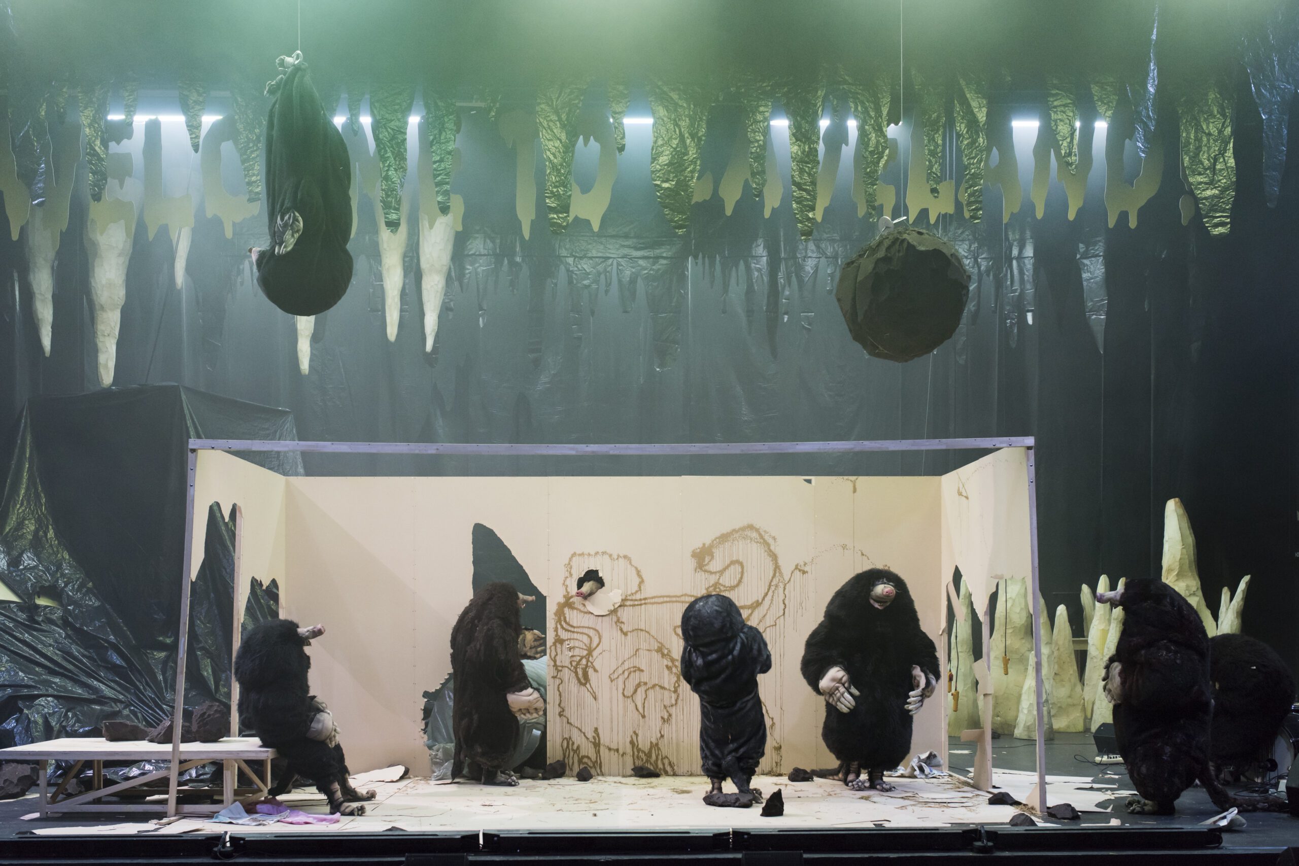 Six people in animal costumes perform onstage in a cave-like set, surrounded by hanging stalactites and a painted wall featuring animal figures. The lighting is dim with a greenish hue.