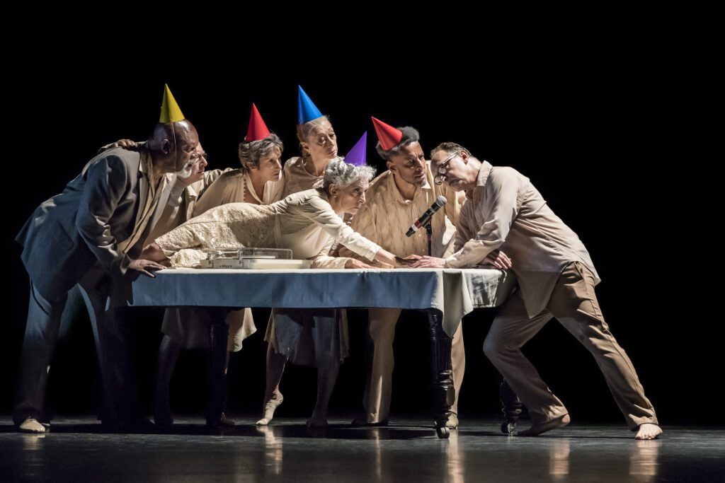 Six adults in party hats gather around a table, leaning in closely while one woman reaches for a cake with a microphone nearby. The scene is on a dark stage, under dramatic lighting.