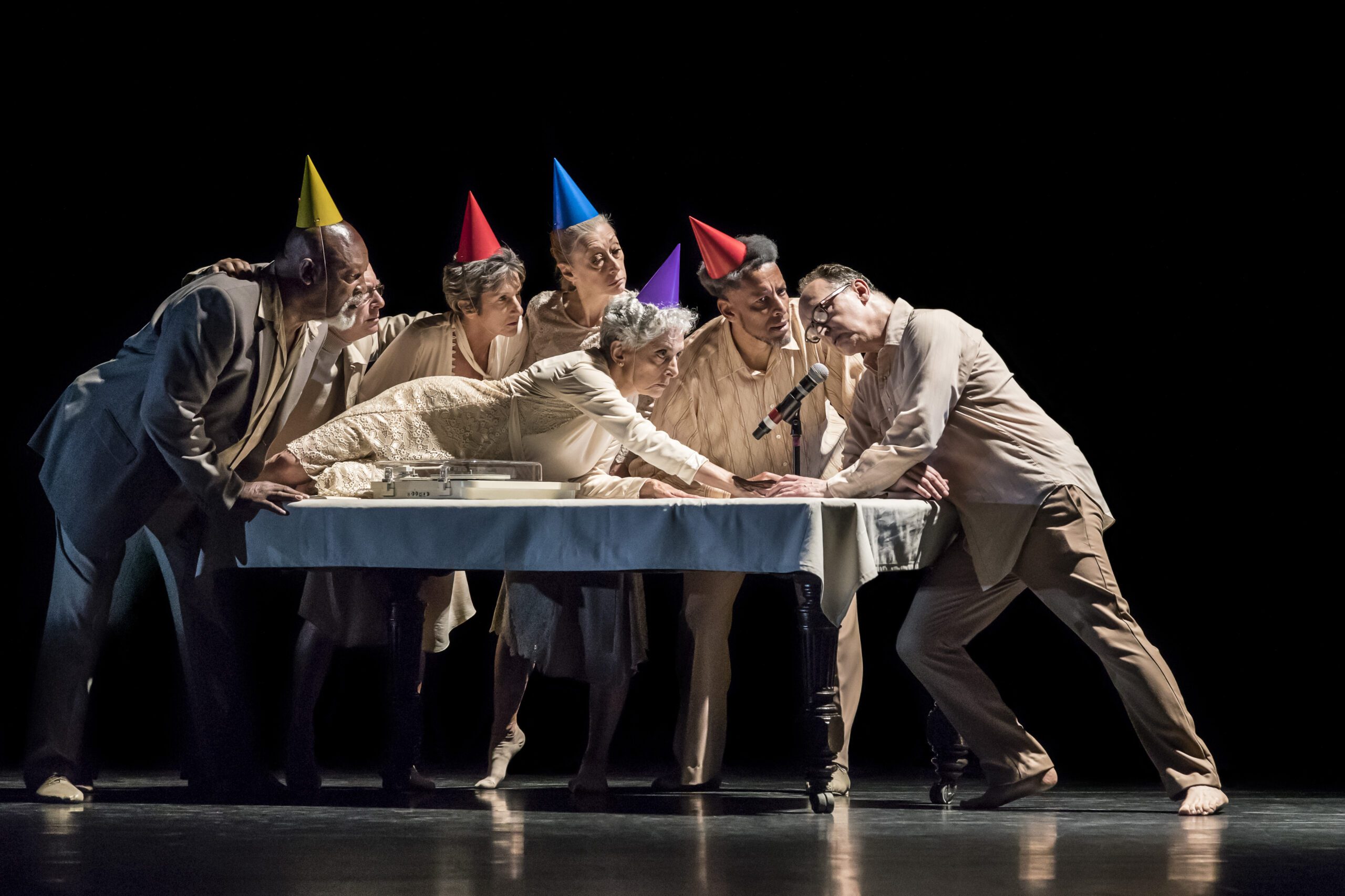 Six adults in party hats gather around a table, leaning in closely while one woman reaches for a cake with a microphone nearby. The scene is on a dark stage, under dramatic lighting.