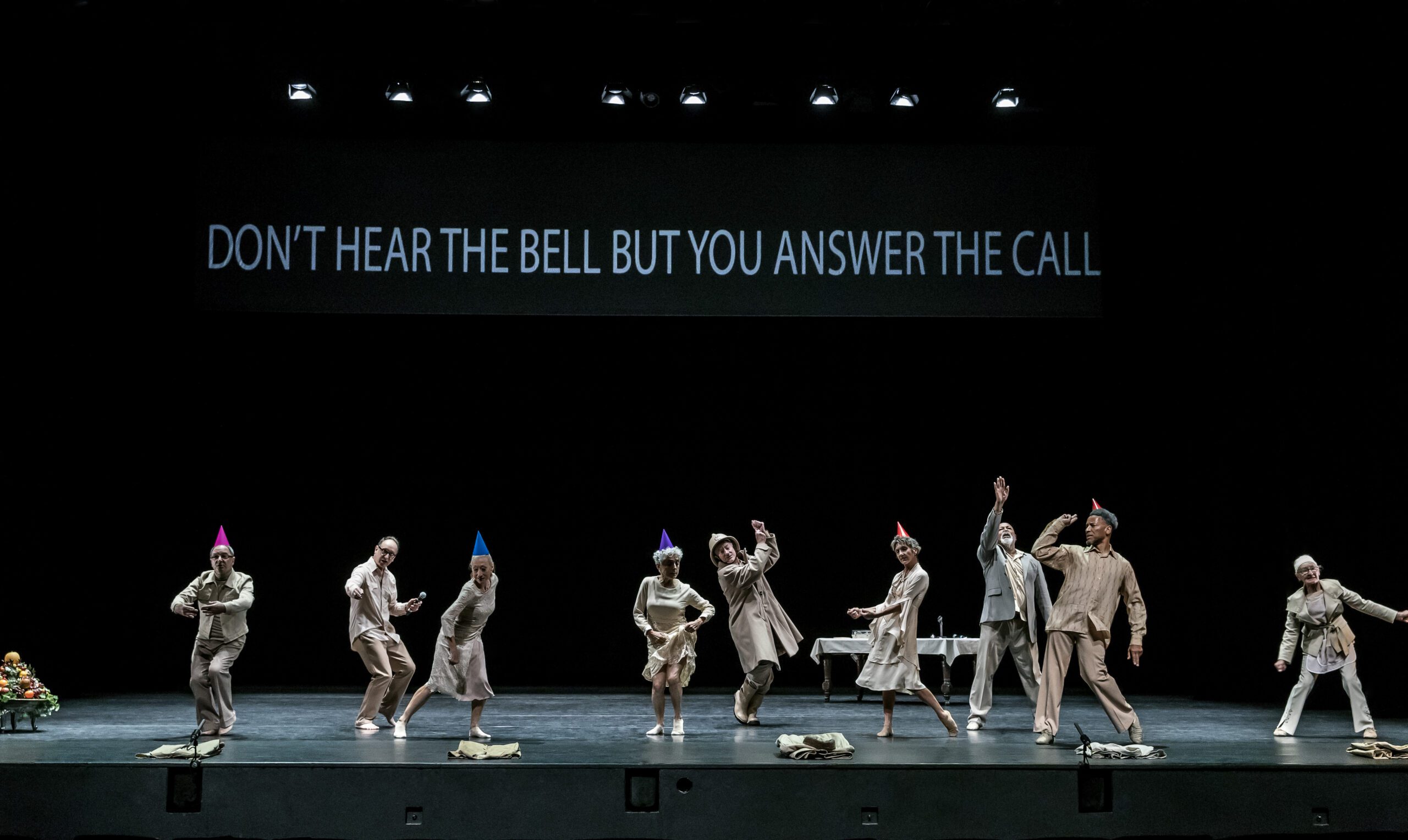 Nine people in party hats dance onstage in muted costumes. Projected above them: “DON’T HEAR THE BELL BUT YOU ANSWER THE CALL.” A table and some objects are in the background.