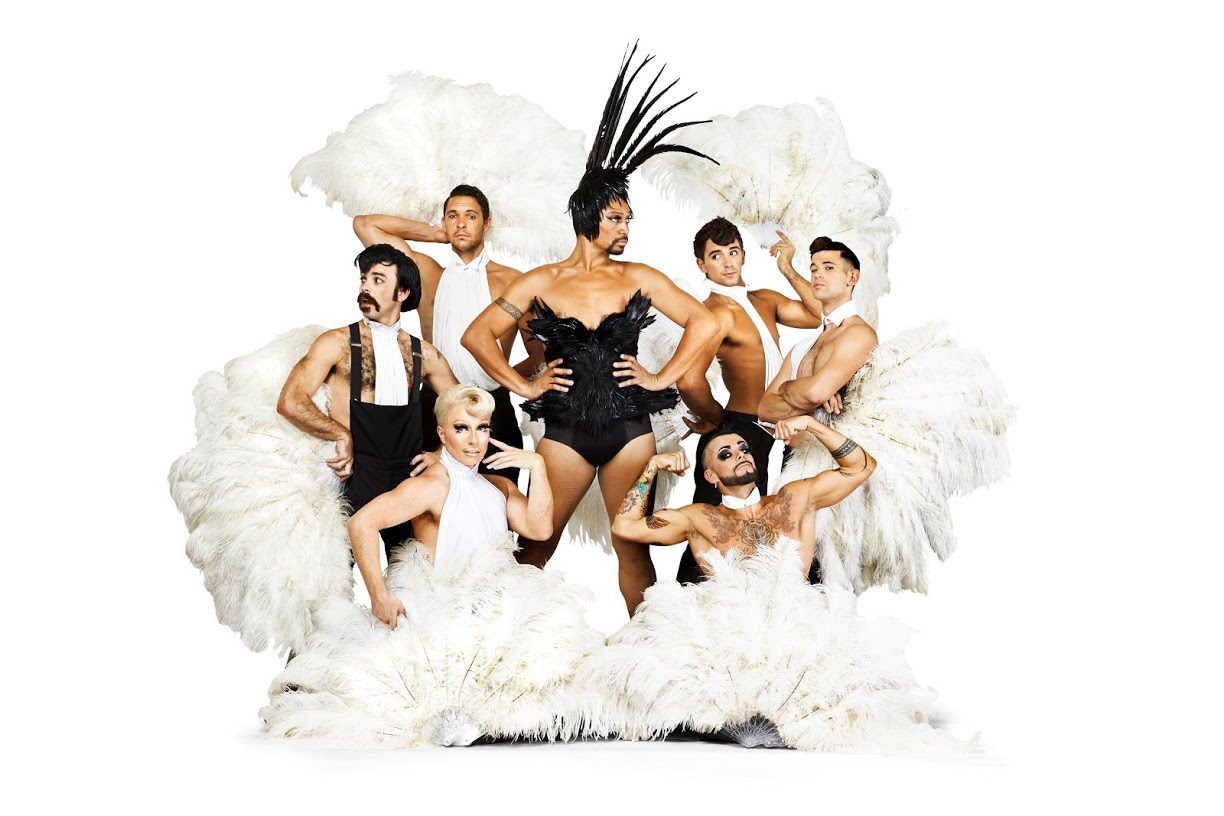 Seven performers pose dramatically with large white feather fans; the central figure wears a black costume and a tall feathered headpiece, while others display expressive poses and facial expressions against a white background.