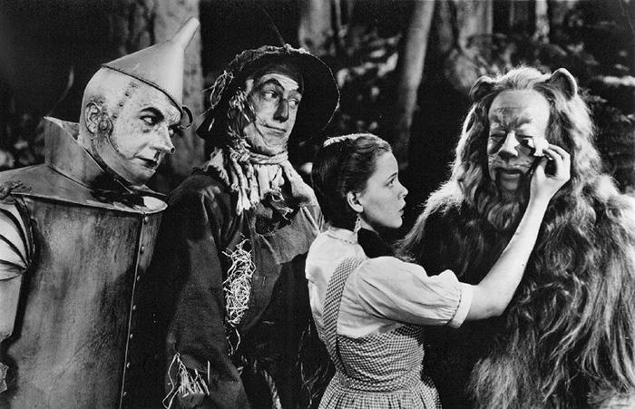 A girl in a gingham dress comforts a lion while standing beside a metal man and a scarecrow in a forest, all in an old black and white film scene.