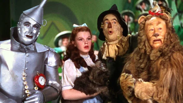 Four characters—Tin Man, Dorothy holding a dog, Scarecrow, and Cowardly Lion—stand close together, looking surprised. They are in colorful costumes on a vibrant set from “The Wizard of Oz.”.