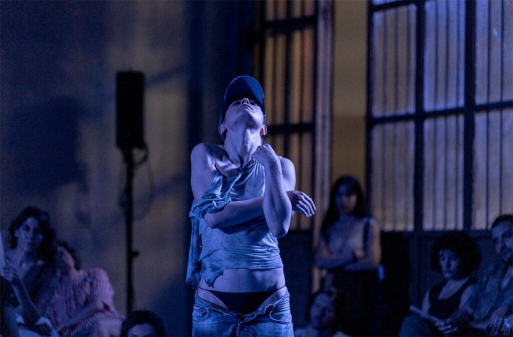 A person in a blue cap and casual clothes performs a dramatic dance under blue lighting, with an audience seated in the background watching attentively.