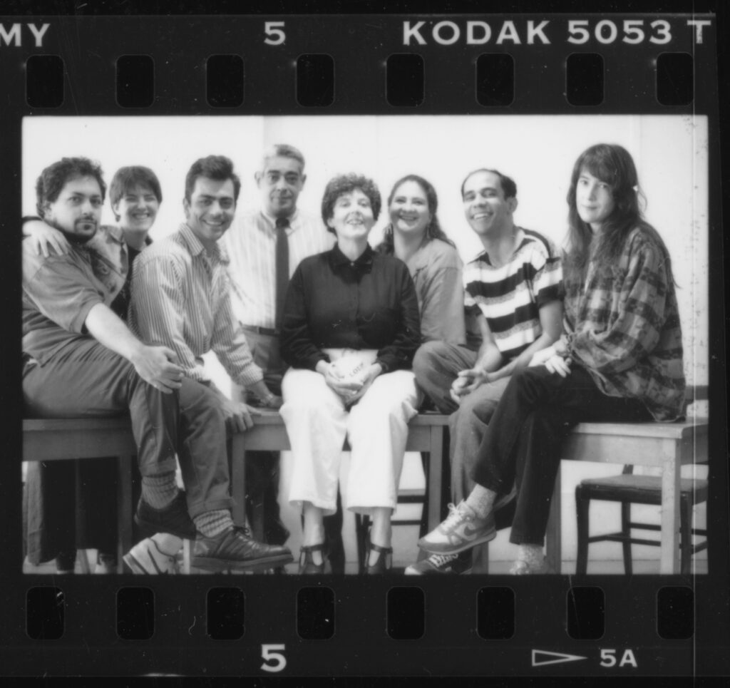 A black-and-white photo of eight people sitting and standing close together, smiling at the camera. The image has a film strip border with “KODAK 5053” text visible at the top.