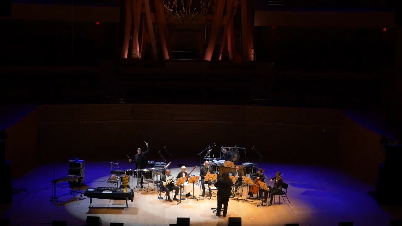 A small orchestra with string and percussion instruments performs on a dimly lit stage, surrounded by blue and purple lighting, with a conductor leading the group.