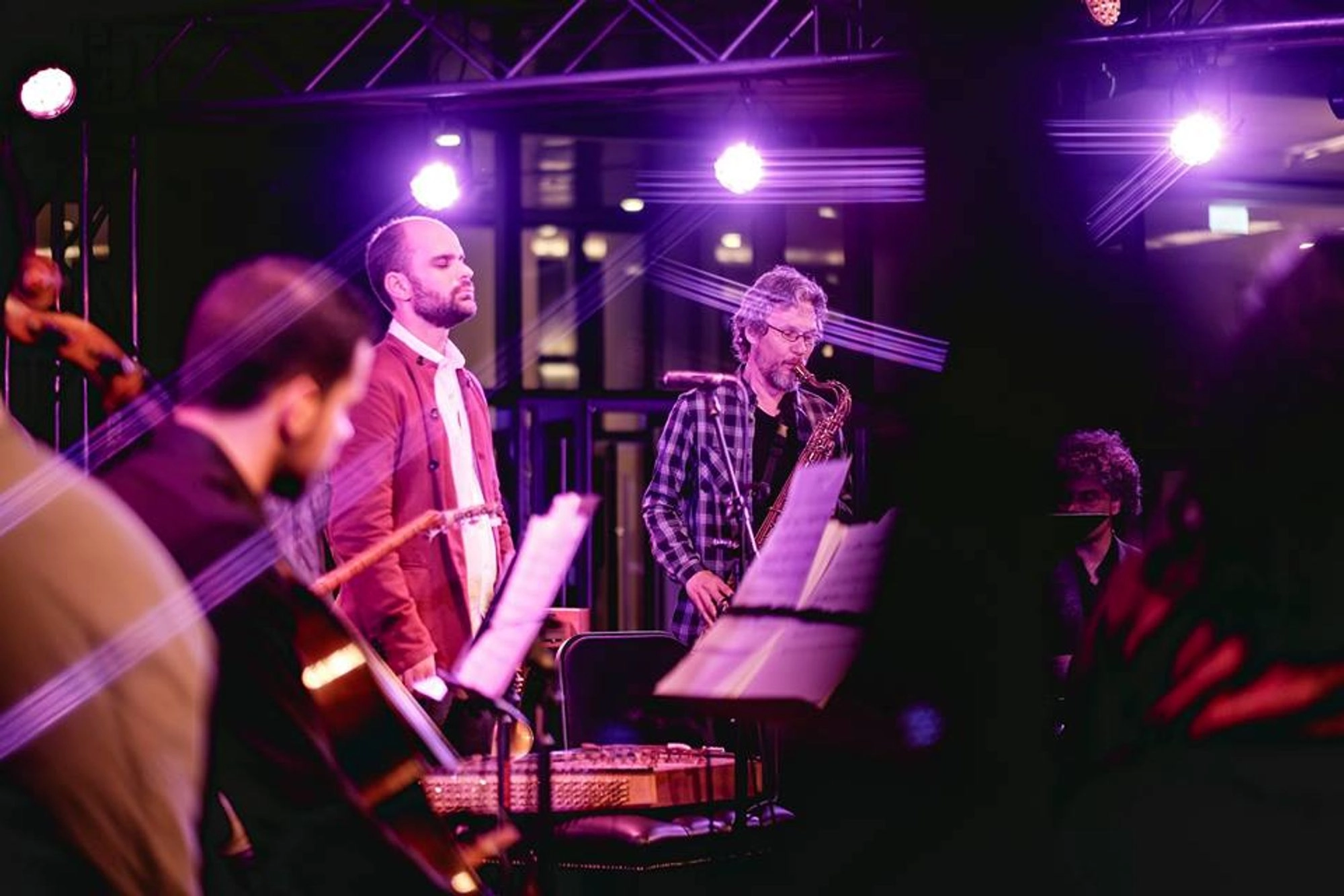 Musicians perform on stage under purple lighting, with music stands and instruments visible. Two men stand in the center, one singing and the other playing a wind instrument. Other musicians are seated in the foreground.
