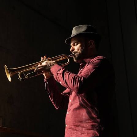 A man in a hat and red shirt plays a trumpet, standing against a dark background with dramatic lighting highlighting his face and instrument.