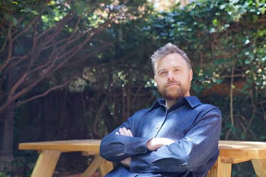 A man with light hair and a beard sits with arms crossed at a wooden picnic table outdoors, surrounded by trees and greenery.
