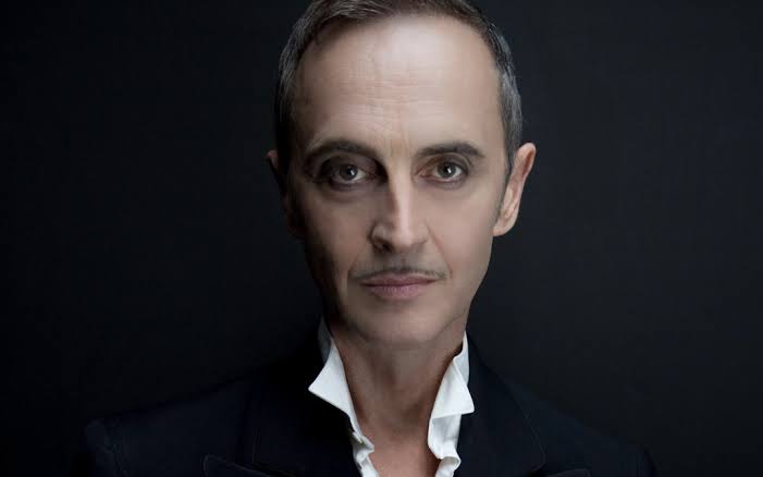 A person with short hair, neatly groomed mustache, and sharp features wears a dark suit with a white ruffled collar, posing against a plain dark background.