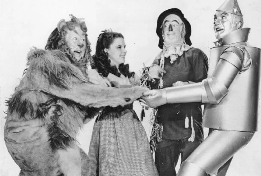 Four characters from “The Wizard of Oz” (the Cowardly Lion, Dorothy, the Scarecrow, and the Tin Man) stand holding hands and smiling at each other against a plain background.
