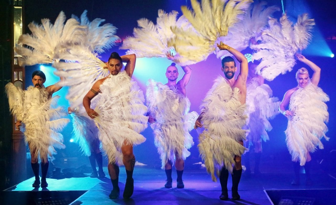 Five performers dressed in feathery white costumes and black boots dance energetically on stage, each holding large feather fans, with blue and purple stage lighting in the background.